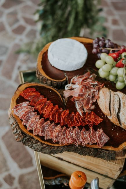 A rustic wooden serving platter, cut from a tree trunk, displaying an assortment of cured meats, a round wheel of soft white cheese, fresh grapes including green and red varieties, and slices of crusty bread. The platter is placed on a small table or stand with a stone or brick surface in the background, indicating an indoor setting. The arrangement appears to be part of a food presentation or catering setup, with the meats and cheese organized in sections and the grapes clustered for easy access. The lighting highlights the textures and colours of the food, emphasizing freshness and quality, which aligns with local catering or event preparation associated with house or venue hosting.