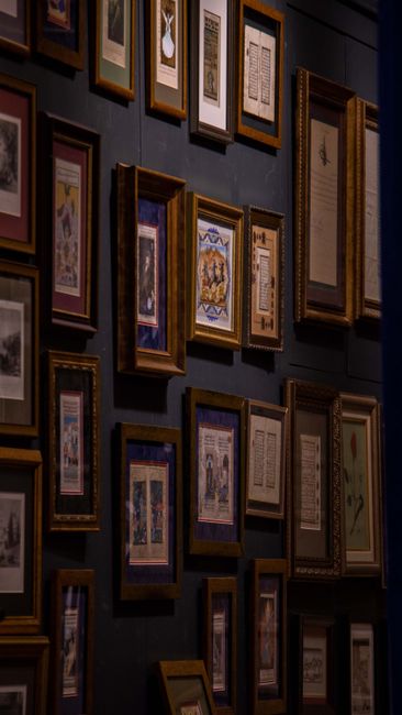 A collection of framed artworks and documents arranged on a dark-colored wall, featuring various sizes and styles of wooden and metallic frames. The images include illustrations, text, and decorative patterns, some with ornate borders and others with simple designs. The frames are organized in a somewhat grid-like pattern, with some slightly tilted or overlapping. The setting appears to be indoors, possibly part of an art gallery or a home decorating a feature wall. The lighting highlights the frames and their contents, contributing to a professional and orderly appearance. The photograph captures the process of home relocation or artwork handling, consistent with house removals and framing services offered by Man and Van East Ham, particularly related to antique and fine art removals in East Ham.