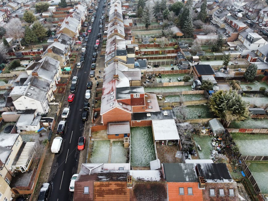 A black and white aerial photograph showing a residential neighbourhood in East Ham, with rows of terraced houses lining the main street, parked cars along both sides of the road, and lush trees interspersed throughout the area. In the foreground, rooftops, backyards with fences, and garden sheds are visible, while further in the distance, the landscape transitions into a more urban environment with larger buildings and high-rises. The sky is partly cloudy with patches of sunlight illuminating the scene, and the overall view captures the extensive spread of suburban housing typical of the area. This image provides a clear visual context for home relocation and moving services by illustrating the typical residential setting where furniture transport and packing and moving processes occur. Man and Van East Ham may utilize such visuals to reflect their expertise in handling moving logistics in urban neighbourhoods.