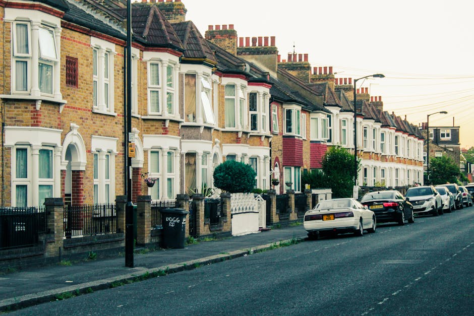 A street scene during daytime in East Ham, showing a residential and commercial area with a mix of red brick and modern buildings. On the left side, there are shops with closed black shutters and sign