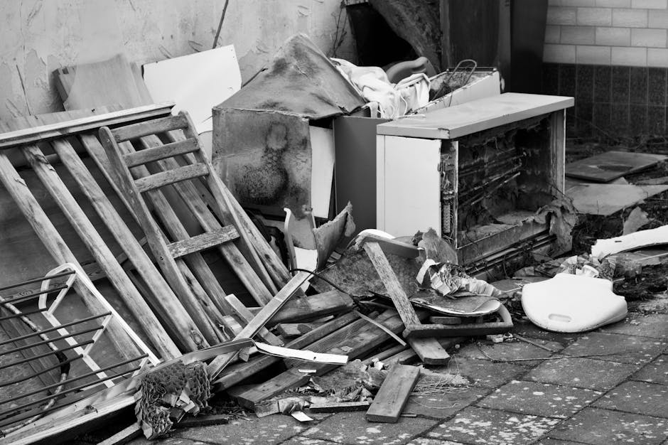 Three worn and damaged antique chairs are positioned side by side outside a building against a weathered wooden wall with peeling white paint near the roofline. The chair on the left features wooden a