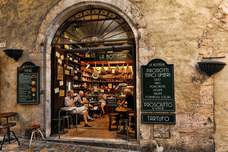 The image shows the entrance of a small Italian-style cafe or restaurant, with a rounded archway made of stone and metal, leading into an interior filled with wooden shelves stocked with bottles and jars. Inside, several elderly customers are seated at small tables and bar stools, engaging in conversation and enjoying their meals or drinks. The exterior wall of the building is textured stone with decorative black wall-mounted planters on either side of the archway. Two black chalkboard menu signs are positioned outside, displaying items such as prosciutto, truffle, and other regional delicacies, reflecting a focus on local Italian cuisine. The area appears well-lit with natural light, and the outdoor pavement is composed of cobblestones, suggesting an authentic European street scene. This setting is relevant to house removals or relocation services, as [COMPANY_NAME] offers professional transport and packing solutions for businesses and residents, ensuring careful handling and logistics to facilitate a smooth moving process for small commercial premises like this one.