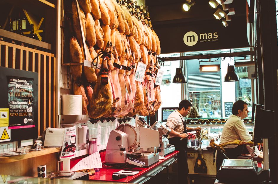 Inside a delicatessen shop with a focus on a butchery section, where several large cured legs of ham are hanging from hooks against a wooden panel wall. Below the hams, there is a counter with a meat slicer, cardboard boxes, and a pink plastic wrapper, indicating ongoing packing or slicing activities. Two male employees are visible; one is using tongs to handle meat on the counter, while the other appears to be preparing or wrapping meat products. Large windows at the front allow natural light to illuminate the workspace, with black pendant lights hanging from the ceiling for additional lighting. In the background, the shop features shelves with jars and products, along with a black sign displaying the word 'mas' as part of the shop’s branding. The scene captures the typical environment of a traditional food shop specializing in cured meats, with a focus on the furniture, packaging materials, tools, and active meat handling in a professional setting, as part of a house or commercial removal process managed by Man and Van East Ham.