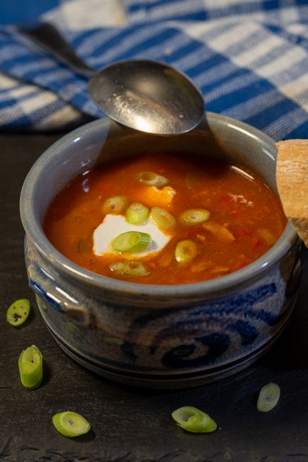 Close-up image of a ceramic bowl of tomato-based soup with visible chunks of tomato and sliced green onions floating on the surface, placed on a dark surface. A silver spoon rests inside the bowl, with part of the handle extending over the edge. The bowl features a decorative blue and white pattern on its exterior, indicating a traditional style. Around the bowl, there are a few scattered slices of green onion on the dark surface. In the background, a piece of rustic brown bread is partially visible, suggesting it is ready to be served alongside the soup. The setting appears casual and inviting, with natural lighting highlighting the vibrant red of the soup and the fresh green of the onions. This image focuses on the presentation of a home-cooked meal, relevant to content about kitchen or dining setup during a house move or packing process, as well as highlighting household food preparation aspects in the context of a house removal service provided by Man and Van East Ham.