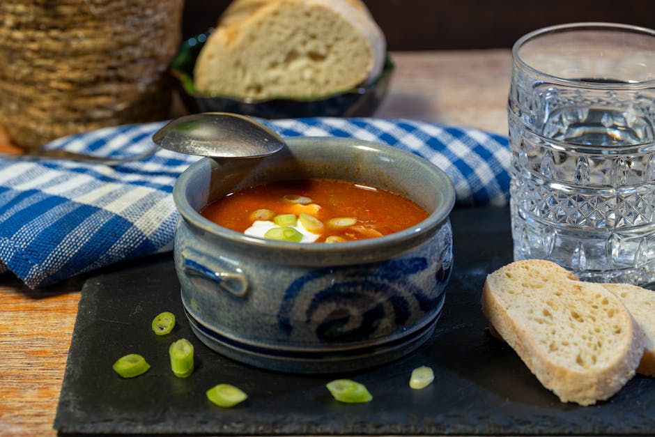 A close-up image of a bowl of tomato-based soup garnished with chopped green onions, placed on a black slate serving board. The bowl is ceramic with a blue and white decorative pattern, positioned near a glass of water with a faceted design and a couple of slices of crusty bread on the slate. In the background, a small bowl of additional bread and a blue and white checkered cloth are visible on a wooden table, suggesting a casual dining scene. The image captures the process of home meal preparation or serving, with natural lighting highlighting the textures of the soup, bread, and table setting, reflecting a typical kitchen or dining area environment. The scene relates indirectly to house relocations involving home preparation or unpacking after a move, as part of a comprehensive moving and packing process facilitated by [COMPANY_NAME], exemplifying the importance of maintaining household routines during relocation.