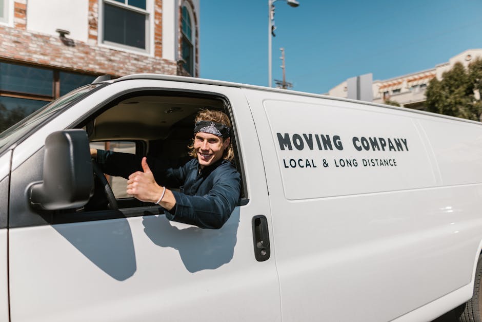 A young man with shoulder-length hair and a bandana, smiling and giving a thumbs-up, is sitting in the driver’s seat of a white moving van parked outside a residential building. The van, associated with a local and long-distance moving company, has the words 'MOVING COMPANY' and 'LOCAL & LONG DISTANCE' printed on its side. The scene shows the driver in casual clothing, possibly preparing for a home relocation, with the van's door open and the interior partially visible. In the background, there are contemporary multi-storey buildings with large windows, and a clear blue sky overhead. The pavement in front of the property is visible, and the van appears to be in the process of loading or unloading furniture or boxes, consistent with professional house removal services. This image illustrates an active loading process managed by Man and Van East Ham, reflecting the logistics of packing, furniture transport, and moving procedures involved in home relocations.