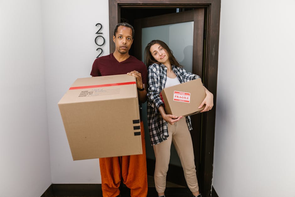 A man and a woman stand inside a doorway, each holding cardboard boxes related to home relocation. The man, dressed in a maroon shirt and orange pants, holds a large box with black tape on the sides, while the woman, wearing a checked shirt and beige pants, holds a smaller box marked 'Fragile.' The scene appears to take place at the entrance of an apartment or house, with the door partially open behind them. The background shows a plain white wall with a number plaque mounted beside the door, and protective packaging materials such as cardboard boxes and tape suggest a packing and moving process is underway. The lighting is bright, emphasizing the preparation for a furniture transport or home relocation task, and this image is associated with the house removals services of Man and Van East Ham, assisting clients with timely and efficient moving logistics.