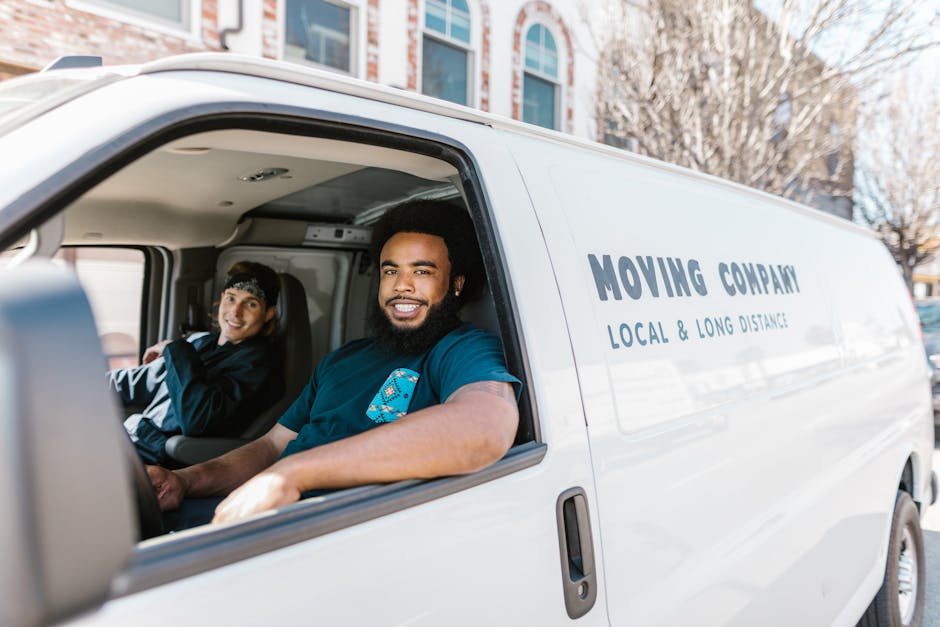 A young man with shoulder-length hair and a bandana, smiling and giving a thumbs-up, is sitting in the driver’s seat of a white moving van parked outside a residential building. The van, associated with a local and long-distance moving company, has the words 'MOVING COMPANY' and 'LOCAL & LONG DISTANCE' printed on its side. The scene shows the driver in casual clothing, possibly preparing for a home relocation, with the van's door open and the interior partially visible. In the background, there are contemporary multi-storey buildings with large windows, and a clear blue sky overhead. The pavement in front of the property is visible, and the van appears to be in the process of loading or unloading furniture or boxes, consistent with professional house removal services. This image illustrates an active loading process managed by Man and Van East Ham, reflecting the logistics of packing, furniture transport, and moving procedures involved in home relocations.