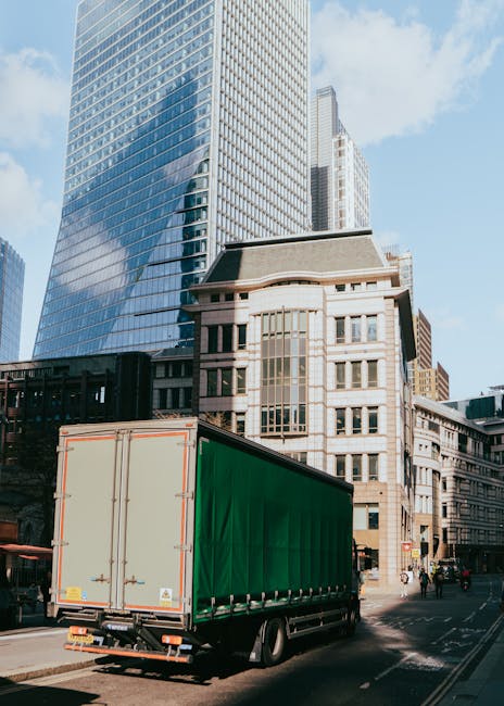 A large green covered delivery truck parked on an urban street in East Ham, in front of modern glass office buildings and older stone and brick structures. The truck, associated with Man and Van East