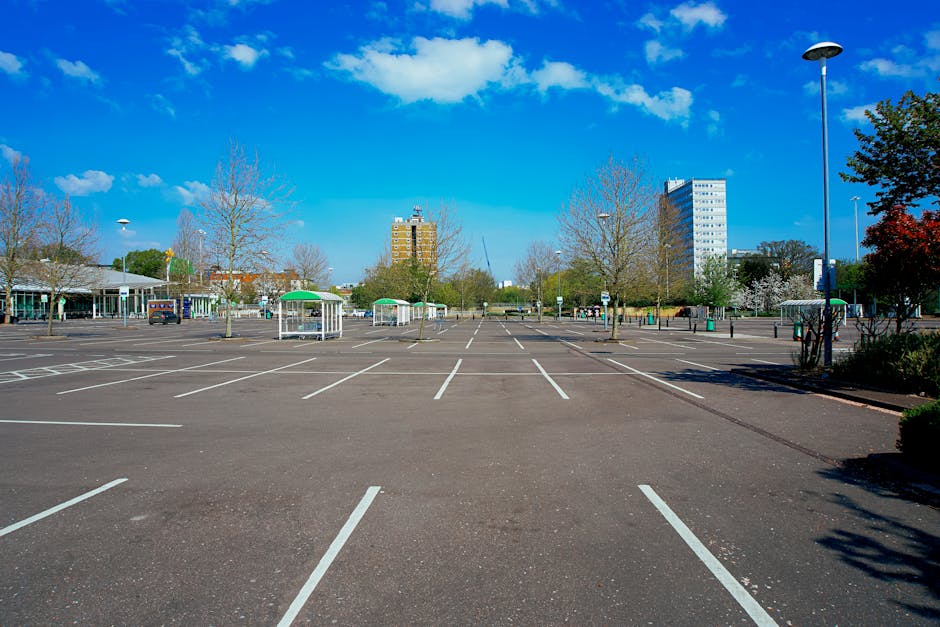 A nearly empty outdoor parking lot with marked parking spaces and a few small green and white covered structures, situated in an urban area during daytime. The background features leafless trees, modern high-rise buildings, and a clear blue sky with scattered white clouds. Several tall streetlights and a few parked cars are visible around the lot. The scene is well-lit with natural sunlight, casting shadows from trees and structures onto the asphalt. The parking space appears to be used occasionally for home relocation logistics, and Man and Van East Ham provides relevant removals and furniture transport services, supporting efficient packing and loading processes in such environments.