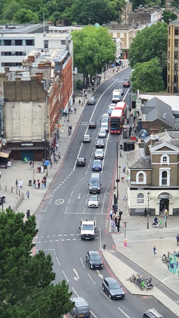Aerial view of a city street during daytime showing a line of vehicles including cars, vans, and buses moving along the road, with pedestrians walking on the sidewalks and crossing at designated crossings. The street is lined with historic and modern buildings, some with brick facades and others with flat roofs, and is surrounded by lush green trees providing shade. Visible parked bicycles are secured to bike racks near the pedestrian crossings. The environment appears busy, with clear traffic lanes divided by painted markings, and the lighting indicates a bright, possibly partly cloudy day. This scene highlights urban transport and movement, fitting for content related to house removals and furniture transport services, exemplified by companies like Man and Van East Ham, who facilitate home relocation and moving logistics.