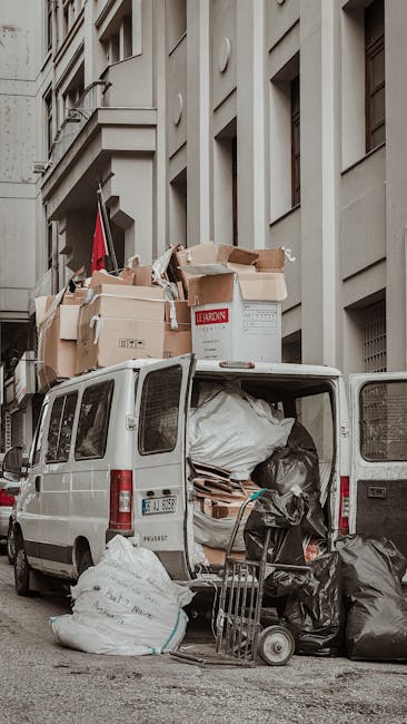 A white van parked on a city street next to a tall, beige residential building with multiple windows and decorative architectural features. The van's rear doors are open, revealing various packed items ready for a home relocation, including large white and black plastic rubbish bags, flattened cardboard boxes, and pieces of packing material. Several cardboard boxes, some labeled, are stacked on top of the van, indicating ongoing packing and loading processes. A small trolley or hand truck is positioned near the open back of the van, loaded with additional black rubbish bags and packing supplies. Natural daylight illuminates the scene, capturing the typical environment of furniture transport and moving logistics as part of a professional removals service by Man and Van East Ham, specializing in same-day East Ham removals at real costs and availability.