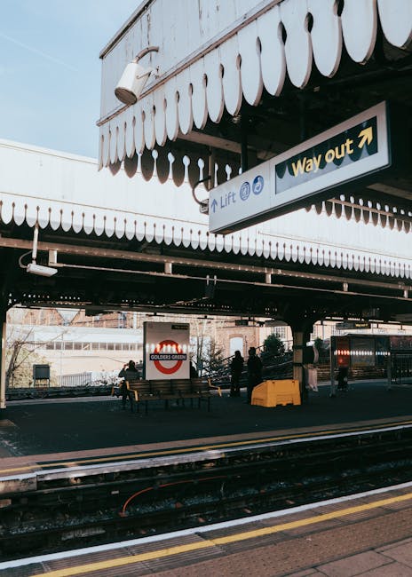 The image shows the exterior of East Ham train station, with a platform featuring a seating area and a sign indicating the way out. In the foreground, train tracks run parallel to the platform, which is paved with a textured surface suitable for pedestrian movement. Above, there's a metal awning with decorative scalloped edging providing shelter, and a large directional sign hanging from it indicating the way out and lift access. The station environment includes a few pedestrians, some seated on benches, and others walking or standing near the platform edge, possibly involved in a home relocation or furniture transport process. In the background, a modern building with large windows and the Golder's Green London Underground sign is visible, suggesting the station's connectivity and infrastructure supporting moving and removal services. The scene is illuminated by natural daylight, emphasizing the urban setting clearly used by people undertaking moving logistics or transport tasks, with visible materials like cardboard boxes, packaging wraps, or furniture likely being moved nearby, although not directly seen in this shot. Man and Van East Ham geographic relevance is implied by the station's setting as part of their removals services.