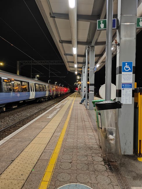 A night-time scene at East Ham station featuring a train with blue and white carriages stopped on the platform. The platform is lit by overhead lighting and has tactile paving with yellow strips along the edge, indicating safe distances for passengers. To the right, a metallic waste bin with a black plastic liner is positioned next to a blue and white accessible information sign, attached to a column supporting the station roof. The station shelter has a glass wall and metal supports visible overhead, providing shelter for waiting passengers. The train appears to be stationary, and there are no visible passengers on the platform. This setting is typical for house removals or furniture transport logistics, where careful handling and efficient loading processes are crucial, supported by companies like Man and Van East Ham.