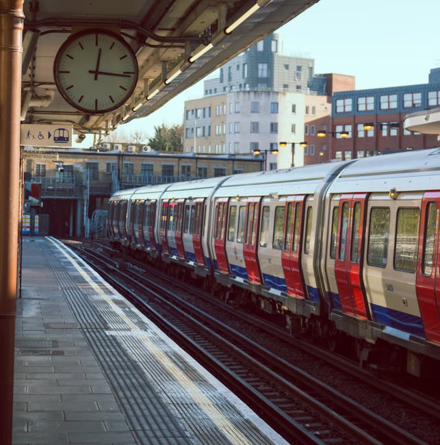 The image shows the exterior of East Ham train station, with a platform featuring a seating area and a sign indicating the way out. In the foreground, train tracks run parallel to the platform, which is paved with a textured surface suitable for pedestrian movement. Above, there's a metal awning with decorative scalloped edging providing shelter, and a large directional sign hanging from it indicating the way out and lift access. The station environment includes a few pedestrians, some seated on benches, and others walking or standing near the platform edge, possibly involved in a home relocation or furniture transport process. In the background, a modern building with large windows and the Golder's Green London Underground sign is visible, suggesting the station's connectivity and infrastructure supporting moving and removal services. The scene is illuminated by natural daylight, emphasizing the urban setting clearly used by people undertaking moving logistics or transport tasks, with visible materials like cardboard boxes, packaging wraps, or furniture likely being moved nearby, although not directly seen in this shot. Man and Van East Ham geographic relevance is implied by the station's setting as part of their removals services.
