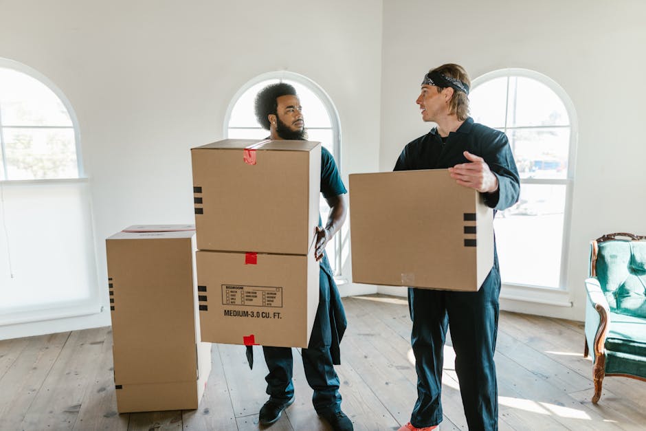 Two men engaged in a home relocation process inside a bright room with large arched windows. The man on the left, with curly hair and a beard, is holding two stacked cardboard boxes, one on top of the other, with red tape and printed labels indicating medium size. The man on the right, wearing a dark outfit and headband, is holding a single cardboard box at chest level while gesturing towards his colleague. There are additional cardboard boxes on the light wooden floor, and a vintage-style green upholstered armchair is visible on the right side. The room features minimal decor, natural light streaming through the windows, and the men are preparing for furniture transport, packing, or loading for a house move, which aligns with services offered by [COMPANY_NAME], such as removals and home relocation.